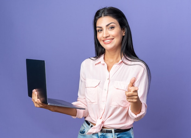 Mooie jongedame in vrijetijdskleding met laptop wijzend met wijsvinger glimlachend vrolijk staande op blauw