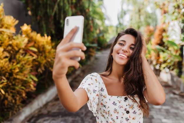 Mooie jongedame in witte zomerblouse met glimlach poseert voor selfie in de tuin