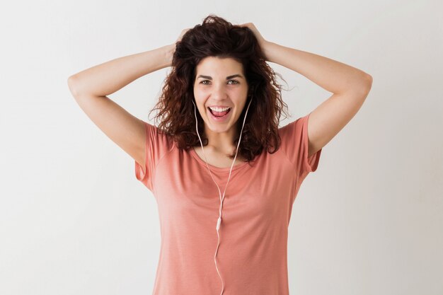 Mooie jongedame luisteren naar muziek op oortelefoons, hand in hand op haar krullend haar, lachen, grappige gezichtsuitdrukking, positieve emotie, gelukkig, geïsoleerd, roze t-shirt, student