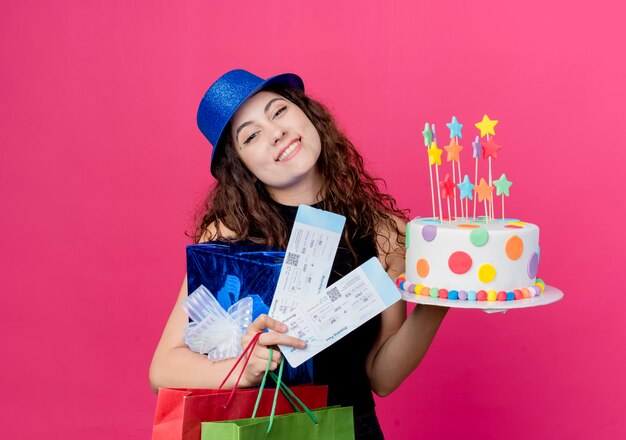 Mooie jongedame met krullend haar in een vakantie hoed bedrijf verjaardagstaart geschenkdoos en vliegtickets blij en blij glimlachend vrolijk verjaardag partij concept over roze