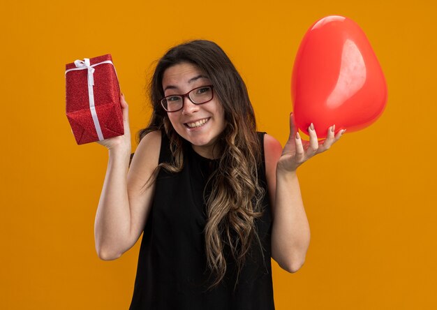 Mooie jongedame met rode ballon in hartvorm en cadeau die er verrast en blij uitziet en vrolijk valentijnsdag viert