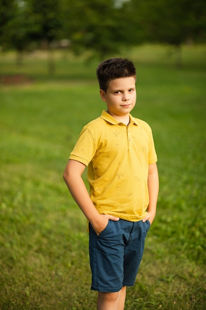 Mooie kleine blanke jongen met donker haar in geel t-shirt en blauwe korte broek die zijn handen in de zakken houdt en glimlacht