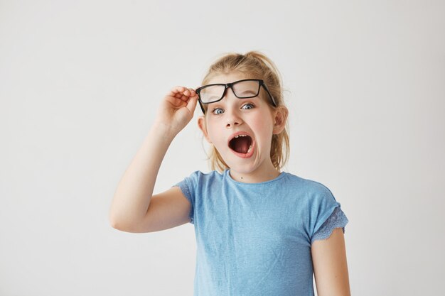 Mooie kleine blonde juffrouw met grote blauwe ogen en licht haar dwaas poseren met wijd geopende mond en het verhogen van haar bril met de hand.