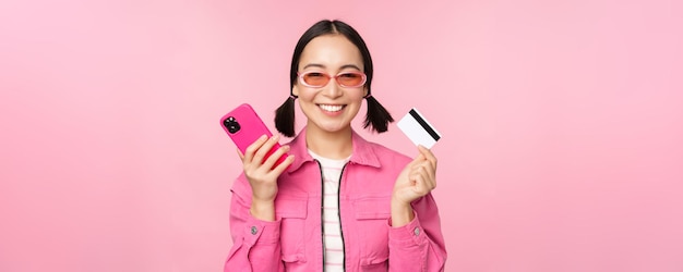 Mooie Koreaanse vrouw met smartphone-creditcard glimlachend naar de camera die online winkelen koopt met een mobiele telefoon die over een roze achtergrond staat