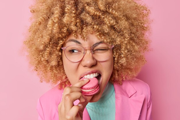 Mooie krullende vrouw bijt hongerige zoete macaron knipoogt ogen kijkt ergens heeft een verslaving aan suiker draagt een bril en formele kleding geïsoleerd over roze achtergrond Heerlijke zoetwaren