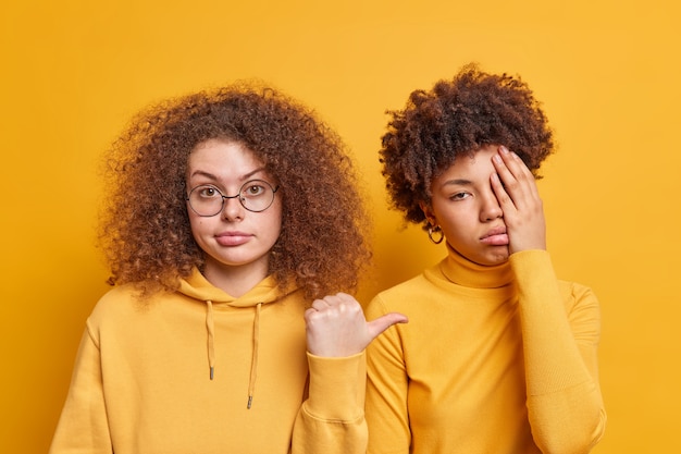 Mooie krullende vrouw wijst met duim naar haar verveelde gefrustreerde vriendin vraagt zich af waarom ze neerslachtig gekleed is, terloops geïsoleerd over gele muur. Mensen emoties en diversiteit concept