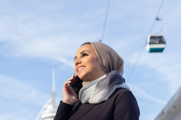 Mooie lachende Arabische vrouw met mobiele telefoon. Vrouw met bedekt hoofd en make-up praten op mobiele telefoon, wegwerp kopje koffie te houden. Internationaal, mooi, social media concept