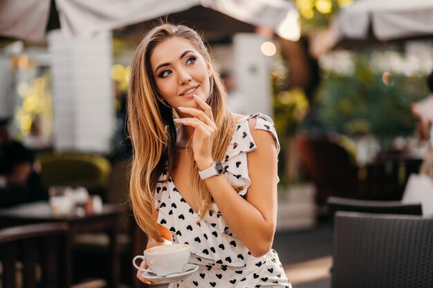 Mooie lachende vrouw, gekleed in stijlvolle witte gedrukte jurk zittend in straat café met kopje cappuccino