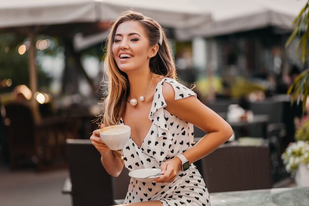Mooie lachende vrouw, gekleed in stijlvolle witte gedrukte jurk zittend in straat café met kopje cappuccino