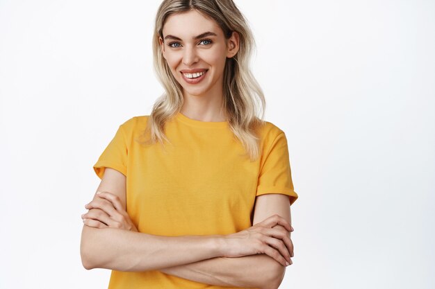Mooie mensen. Jong blond meisje ziet er zelfverzekerd en glimlachend uit, draagt een geel t-shirt, kruisarmen op de borst als een professional, staande op wit.