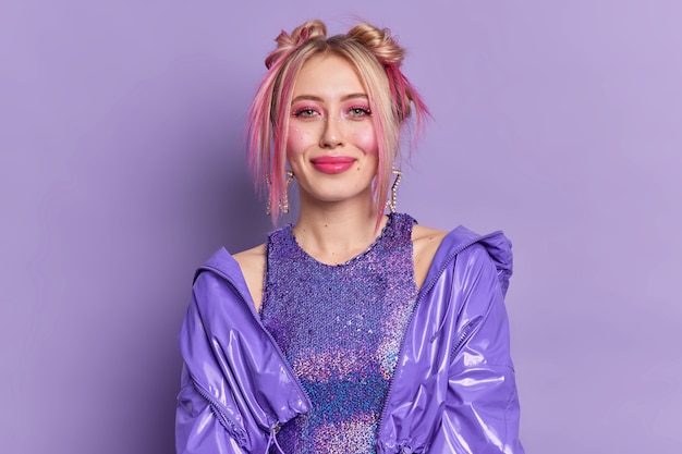 Mooie modieuze blonde vrouw met vrolijke uitdrukking draagt professionele make-up heeft twee haarbroodjes gekleed in stijlvolle kleding poses tegen levendige paarse muurjurken voor discofeest.