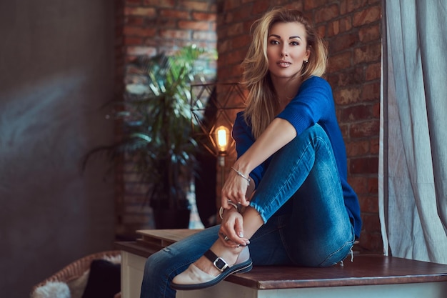 Mooie modieuze blonde vrouw zittend op een tafel tegen een bakstenen muur in een studio met een loft-interieur.