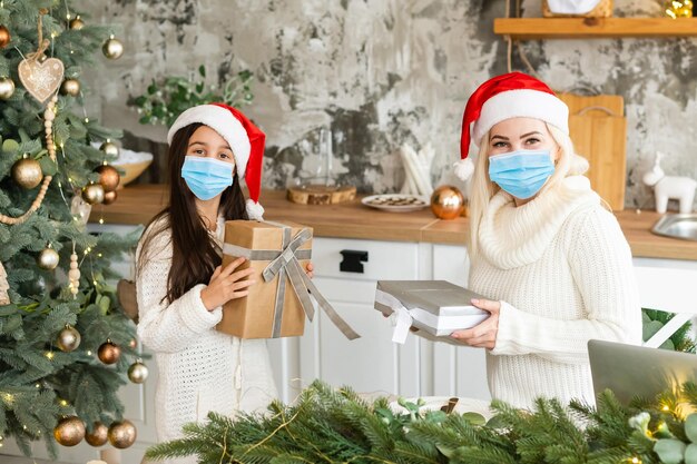 Mooie moeder en dochter met medische maskers hebben plezier thuis bij de kerstboom in een interieur. Gezinsgeluk, vakantie, vreugde, vakantie, spelletjes met een vrouw. Voorbereidingen voor het nieuwe jaar.