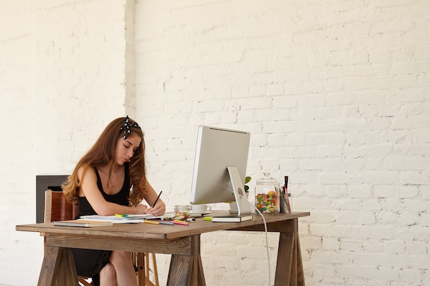 Mooie mooie jonge vrouw die zwarte kantoorjurk en bw-hoofddoek draagt die aan bureau met PC-computer zit terwijl het maken van reclame voor privékleuterschool. Creativiteit, kunst, zaken, werkconcept