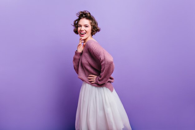 Mooie opgewonden vrouw met stijlvol kapsel poseren in lange witte rok. Binnenfoto van vrolijk donkerbruin vrouwelijk model dat op heldere purpere muur wordt geïsoleerd.