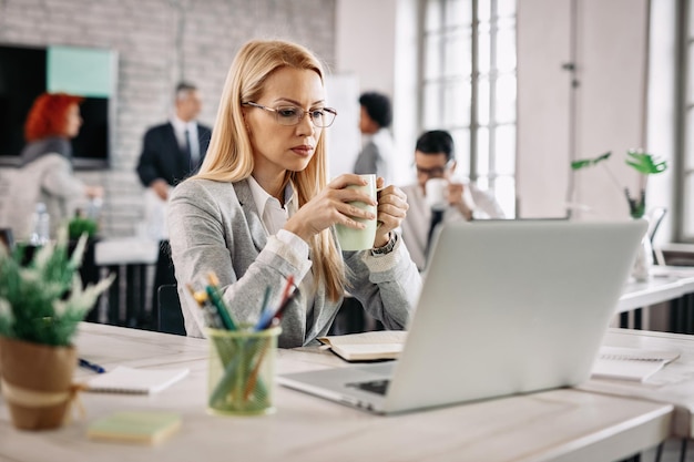 Mooie peinzende zakenvrouw die iets op laptop leest en thee drinkt terwijl ze aan haar bureau op kantoor zit Er zijn mensen op de achtergrond