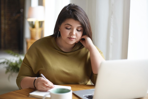 Mooie plus grootte jonge vrouwelijke blogger die in haar dagboek opschrijft, inhoudstrategie voor haar blog ontwikkelt, aan bureau voor open laptop zit, koffie drinkt. Modern technologieconcept