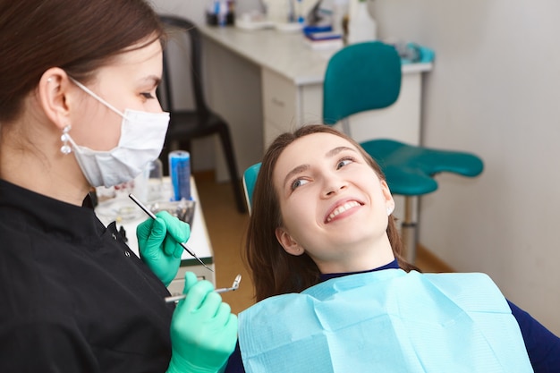 Mooie positieve jonge vrouw die breed glimlacht na regelmatige tandcontrole, die haar vrouwelijke hygiënist bekijkt, haar perfecte witte tanden toont