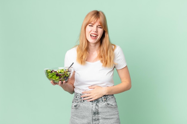 Mooie roodharige vrouw die hardop lacht om een of andere hilarische grap en een salade vasthoudt. dieet concept