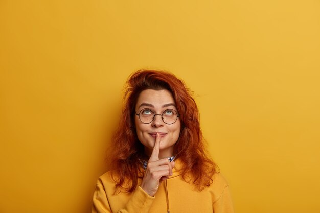 Gratis foto mooie roodharige vrouw kijkt naar boven, houdt de wijsvinger over de lippen, heeft een mysterieuze geheime blik boven