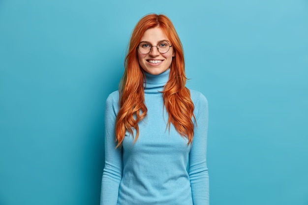Mooie roodharige vrouw met perfecte witte tanden die in een goed humeur is, geniet van prettige gesprekken, kijkt in verrukking en draagt een casual coltrui.