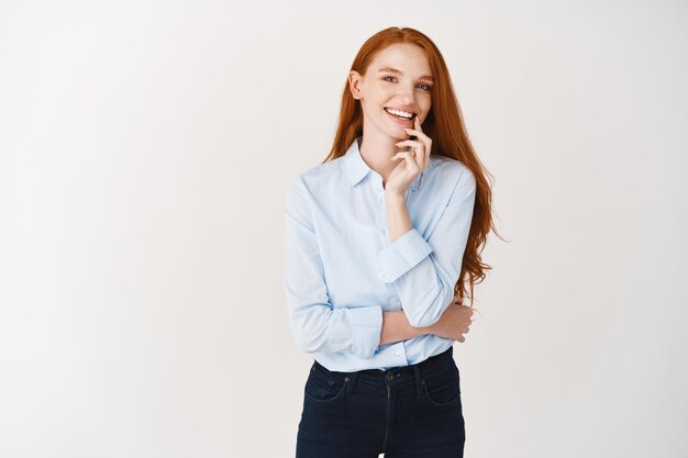 Mooie roodharige zakenvrouw die er gelukkig uitziet en naar voren glimlacht, poserend in een blauw shirt en spijkerbroek