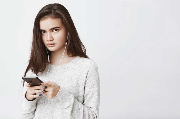 mooie schattige Europese vrouw met lang bruin haar, smartphone te houden terwijl u luistert naar muziek in de koptelefoon, uiting geven aan bezorgdheid.