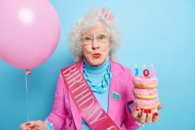 Mooie senior vrouw met krullend haar houdt lippen gevouwen geniet van verjaardagsfeest houdt stapel heerlijke donuts met kaarsen opgeblazen ballon