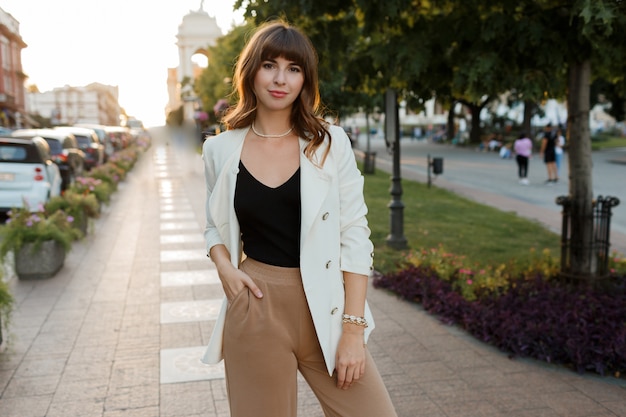 Mooie sexy brunette meisje in casual kleding met perfecte figuur wandelen rond het stadscentrum. elegante stijl. witte jas.