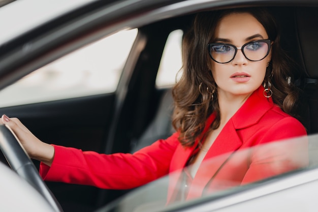 Mooie sexy rijke zakenvrouw in rood pak zit in witte auto, bril