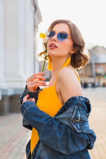Mooie sexy stijlvolle vrouw in gele stijlvolle jurk dragen spijkerjasje, trendy outfit, lente zomer modetrend, zonnige, blauwe zonnebril, straatmode, hipster stijl, modieuze accessoires