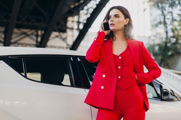 Mooie sexy vrouw in rood pak poseren bij auto praten over zaken op telefoon