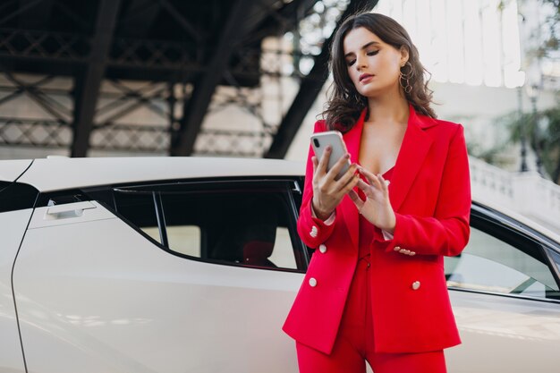 Mooie sexy vrouw in rood pak poseren bij auto praten over zaken op telefoon