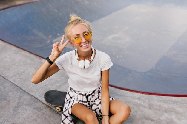 Mooie skater vrouw met trendy kapsel poseren met vredesteken. modieuze gelooide dame in koptelefoon zittend op skateboard.