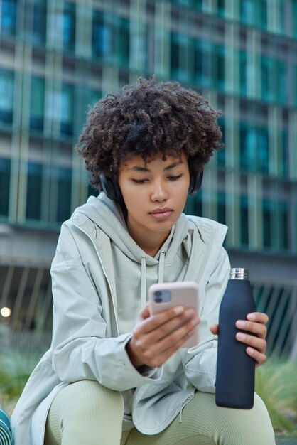 Mooie sportieve vrouw gekleed in sportkleding drinkt zoet water heeft pauze na training luistert favoriete afspeellijst via koptelefoon chats online houdt mobiele telefoon poses tegen stadsgebouw.