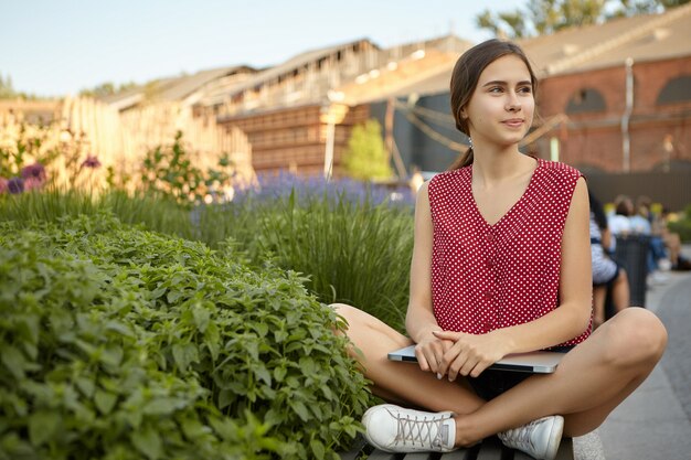 Mooie stijlvolle jonge vrouw freelancer dragen witte sneakers en rode polka top zittend op een bankje benen gekruist houden, laptop vasthouden, met behulp van gratis wifi in stadspark voor verre werk op afstand