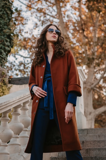 Mooie stijlvolle lachende magere vrouw met krullend haar lopen in straat trappen gekleed in warme bruine jas en blauw pak, herfst trendy mode Streetstyle