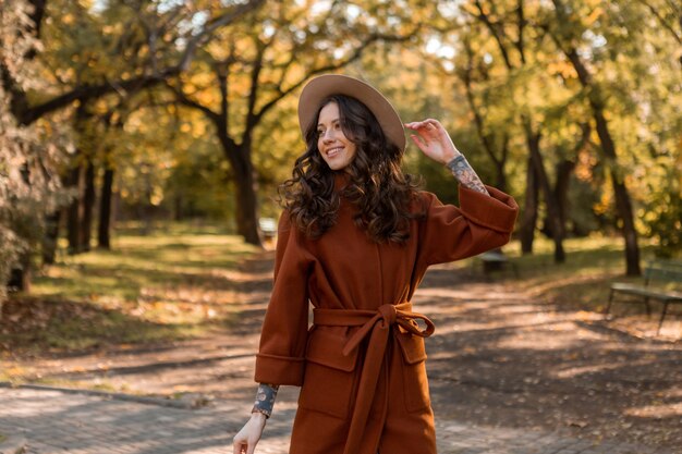 Mooie stijlvolle lachende magere vrouw met krullend haar wandelen in het park gekleed in warme bruine jas, herfst trendy mode Streetstyle