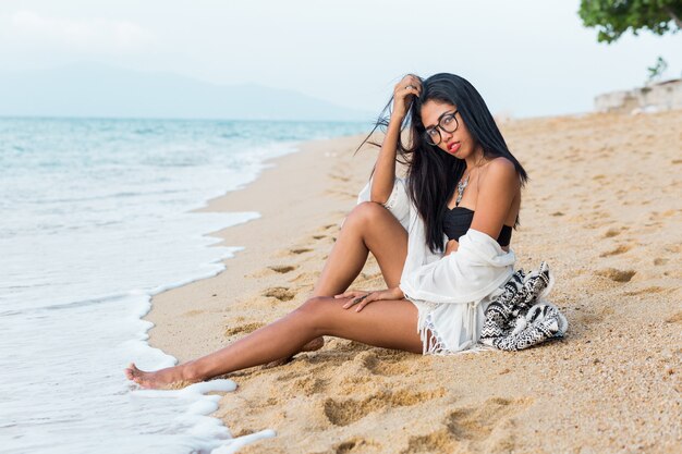Mooie tan Aziatische vrouw met rode lippen zittend op verzenden in de buurt van zee in witte strand Boheemse outfit Stijlvolle vrouw rustend op tropisch strand Vakantie en vakantie concept