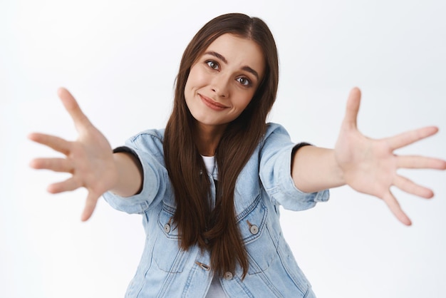 Gratis foto mooie tedere en romantische brunette vriendin in spijkerjasje tilt hoofd schattig uitrekkende handen naar camera om te knuffelen die iemand wil knuffelen en kussen die op een witte achtergrond staat