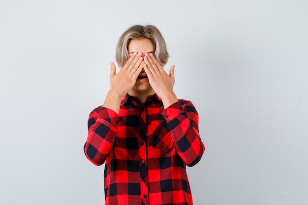 Gratis foto mooie tienerjongen met handen op ogen in geruit overhemd en opgewonden, vooraanzicht.