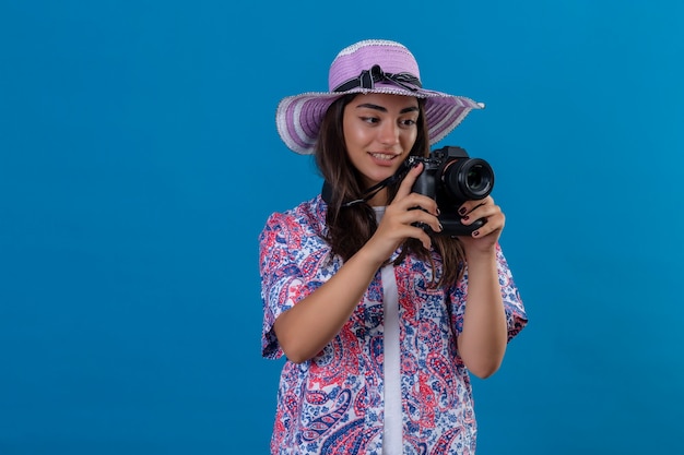 Mooie toeristenvrouw in de zomerhoed met fotocamera met glimlach op gezicht positief en gelukkig status over geïsoleerde blauwe ruimte