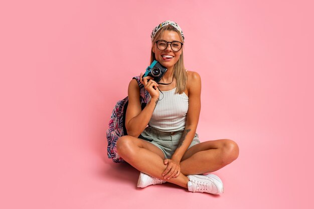 Mooie toeristische vrouw met camera en rugzak zittend op de vloer in studio op roze achtergrond Vakantiestemming