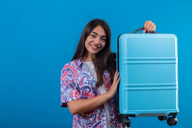 Mooie toeristische vrouw met reiskoffer glimlachend vrolijk klaar voor vakantie staande over geïsoleerde blauwe ruimte