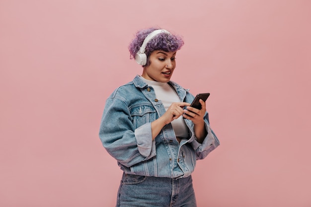 Gratis foto mooie volwassen vrouw met kort haar in een oversized jas en spijkerbroek schrijft sms. vrouw in witte koptelefoon poseren met telefoon op roze.