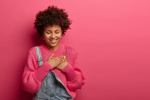 Mooie vrolijke Afro-Amerikaanse vrouw drukt de handpalmen naar het hart, heeft een dankbare uitdrukking