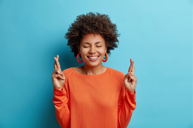 Mooie vrolijke jonge vrouw met brede perfecte glimlach houdt vingers gekruist gelooft in geluk draagt oranje trui geïsoleerd over blauwe muur