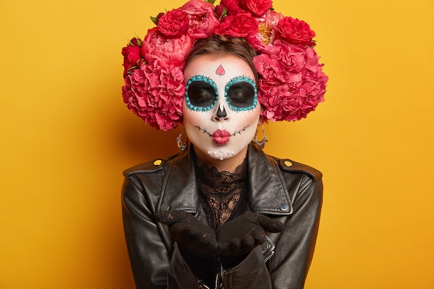 mooie vrouw blaast mwah, houdt lippen gevouwen, draagt creatieve make-up, bereidt zich voor op carnaval, bereidt zich voor op Day of Dead