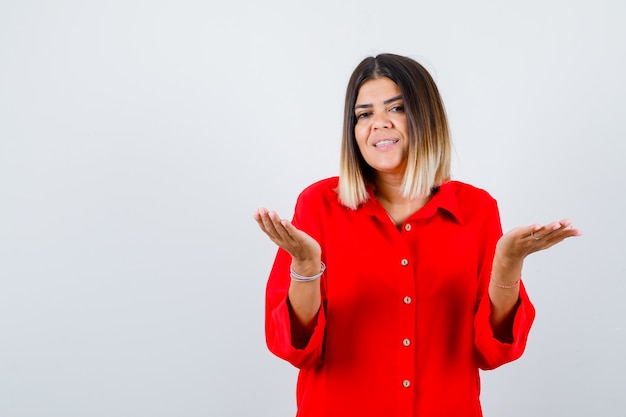 Mooie vrouw die een vraaggebaar maakt in een rode blouse en er vrolijk uitziet, vooraanzicht.