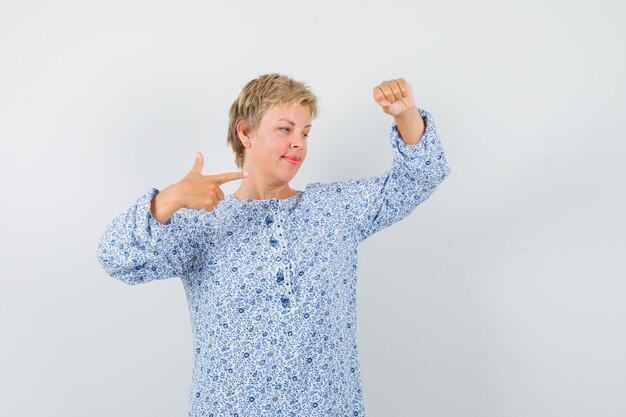 Mooie vrouw die zich voordeed als iets optillen terwijl ze ernaar wijst in een blouse met patroon en er gefocust uitziet. vooraanzicht.